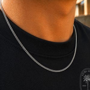 May include: A thin silver-colored curb chain necklace resting against a black t-shirt. The chain features small, uniform interlocking links that create a smooth, continuous metallic line around the neck area. The metal has a polished finish.