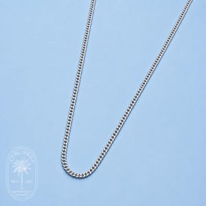 以下が含まれることがあります： 水色の背景に、シルバーのカーブチェーンネックレス。チェーンは、連動するリンクで構成され、テクスチャのある表面を作り出しています。ネックレスは斜めに配置されています。画像は、クリーンでミニマリストな美学を持っています。