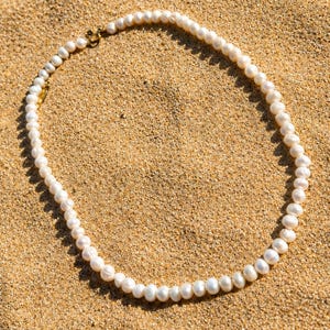 May include: A delicate pearl necklace with a gold clasp rests on a bed of sand. The necklace features numerous white pearls, each with a slightly irregular shape. The gold clasp adds a touch of elegance to the natural beauty of the pearls.