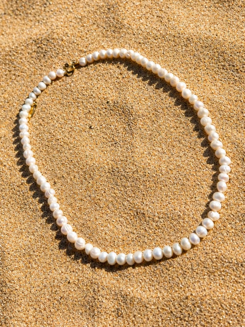 May include: A delicate pearl necklace with a gold clasp rests on a bed of sand. The necklace features numerous white pearls, each with a slightly irregular shape. The gold clasp adds a touch of elegance to the natural beauty of the pearls.