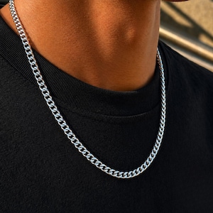 May include: A polished silver-tone curb chain necklace featuring interlocking flat, oval-shaped links. The metallic chain rests against a plain black fabric surface, showcasing a reflective, smooth finish throughout the entire length of the jewelry piece.