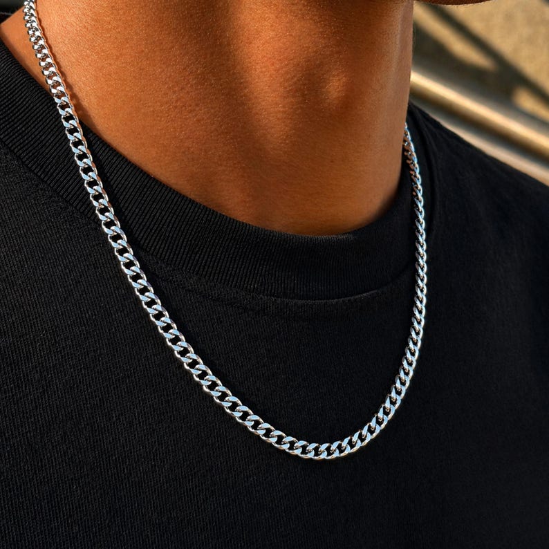 May include: A polished silver-tone curb chain necklace featuring interlocking flat, oval-shaped links. The metallic chain rests against a plain black fabric surface, showcasing a reflective, smooth finish throughout the entire length of the jewelry piece.