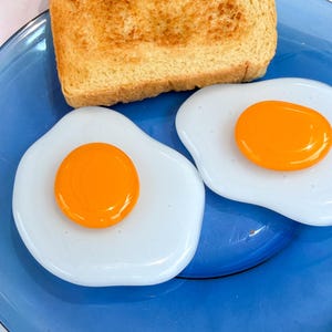 May include: A translucent blue glass plate holding two fried eggs with bright orange yolks and white egg whites, accompanied by a single slice of golden-brown toasted bread. The items are arranged on the circular plate with a smooth finish.