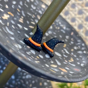 May include: Two black witch hat earrings with orange trim. The earrings are on a black and white speckled surface.