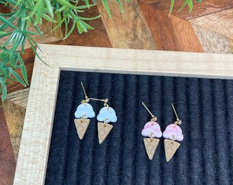 May include: A pair of ice cream cone earrings. One earring has a light blue ice cream scoop and the other has a pink and white ice cream scoop. Both scoops are topped with a brown waffle cone.