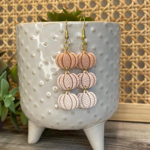 May include: A pair of dangle earrings featuring three stacked peach-colored pumpkin charms with a gold-tone finish.