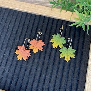 May include: A pair of gold hoop earrings with autumn leaf charms. The leaves are made of polymer clay and are painted in shades of orange, yellow, and green.