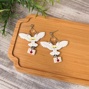 May include: A pair of dangle earrings featuring white owls with yellow eyes and black details, each carrying a small white envelope with a red wax seal. The owls are attached to silver-toned metal posts with circular connectors and textured wings.