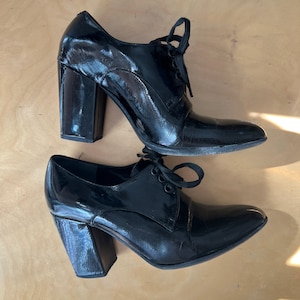 Può includere: Coppia di scarpe con tacco in vernice nera con lacci. Le scarpe hanno un tacco a blocco spesso e una punta a punta.