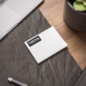May include: A stack of white sticky notes with the word "THINK" in a black rectangle. The notes are on a dark gray surface, with a laptop and pen visible in the background. A potted plant is also in the background.