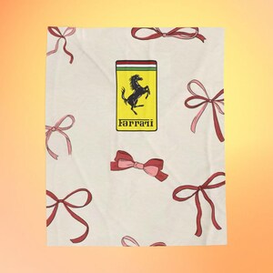 May include: A cream-colored textile featuring a pattern of red and pink bows. The Ferrari logo, a black horse on a yellow background, is centered. The bows vary in size and design, adding a playful touch to the fabric.