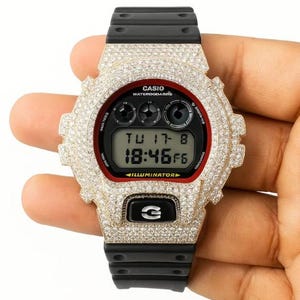 Op de afbeelding: Een Casio G-Shock horloge met een zwarte band en een bezel bezet met sprankelende stenen. De wijzerplaat is zwart met een rode rand, met de tijd en datum. Het woord "ILLUMINATOR" is zichtbaar.