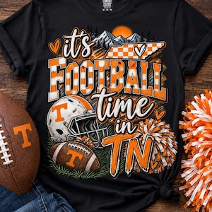 Op de afbeelding: Een zwart t-shirt met een afbeelding met de tekst 'it's FOOTBALL time in TN'. Het ontwerp bevat een footballhelm, een football, een kaart van Tennessee met een ruitpatroon, bergen, een zon en oranje en witte pompons.