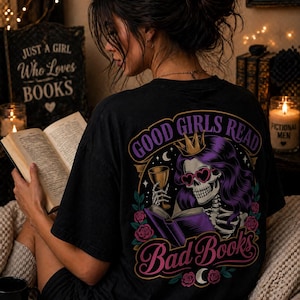 May include: Black t-shirt with a graphic on the back that reads "GOOD GIRLS READ Bad Books". The graphic features a skeleton with purple hair, heart-shaped sunglasses, and a crown, holding a glass and a book. A mug with "EMOTIONAL DAMAGE" is visible.