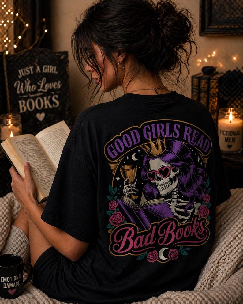May include: Black t-shirt with a graphic on the back that reads "GOOD GIRLS READ Bad Books". The graphic features a skeleton with purple hair, heart-shaped sunglasses, and a crown, holding a glass and a book. A mug with "EMOTIONAL DAMAGE" is visible.