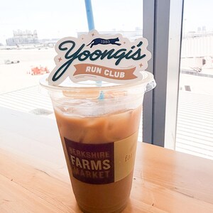 May include: A clear plastic cup filled with iced coffee, featuring a tan sleeve labeled Berkshire Farms Market. A light blue straw protrudes from the top, adorned with a white decorative topper that reads Yoongi's Run Club 1993 in dark green and tan.