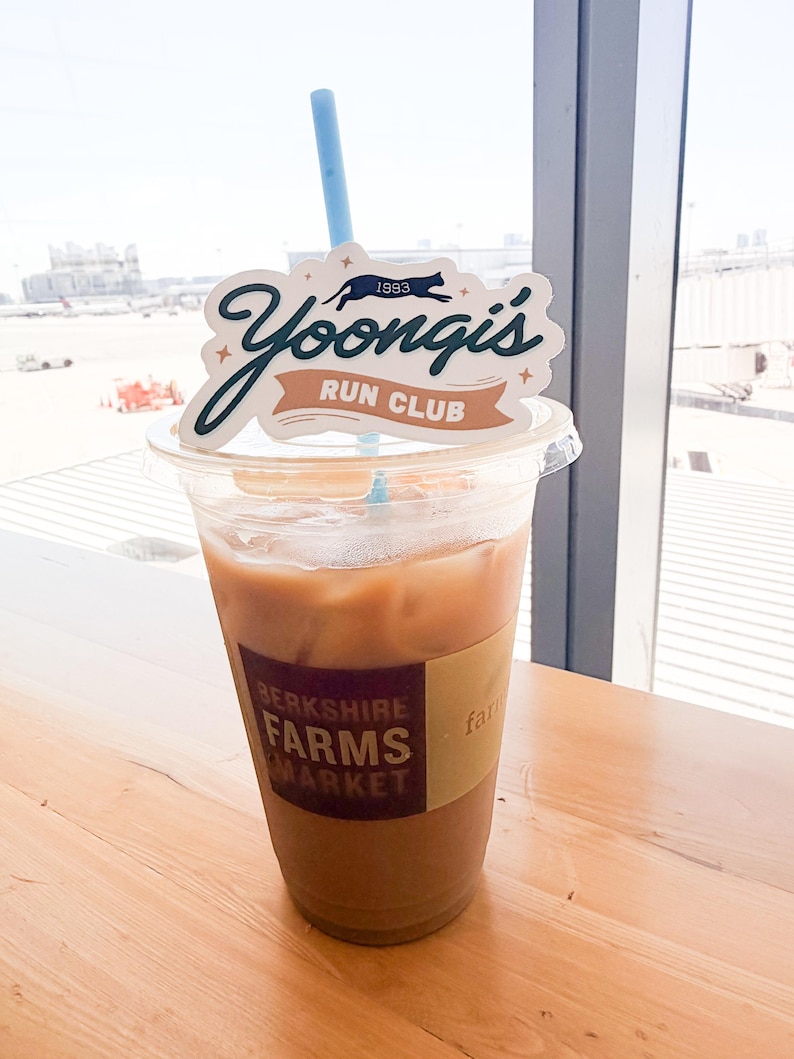 May include: A clear plastic cup filled with iced coffee, featuring a tan sleeve labeled Berkshire Farms Market. A light blue straw protrudes from the top, adorned with a white decorative topper that reads Yoongi's Run Club 1993 in dark green and tan.