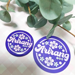 Peut inclure: Deux autocollants ronds violets avec le mot "Arirang" en écriture blanche, entourés de motifs floraux et géométriques blancs. L'un est un bouton, l'autre un autocollant plus grand. Des feuilles vertes sont en arrière-plan.