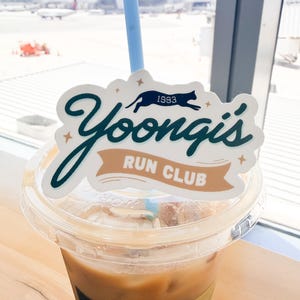 May include: A clear plastic iced coffee cup with a light blue straw and a decorative sticker attached. The sticker features the dark green text Yoongi's, a black cat silhouette, the year 1993, and a tan banner reading RUN CLUB with small stars.