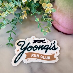 May include: A die-cut sticker featuring the black text Yoongi's in a cursive font, with the words RUN CLUB in white on a tan banner. Above the text is a black silhouette of a running cat with the number 1993, accented by small four-pointed stars.