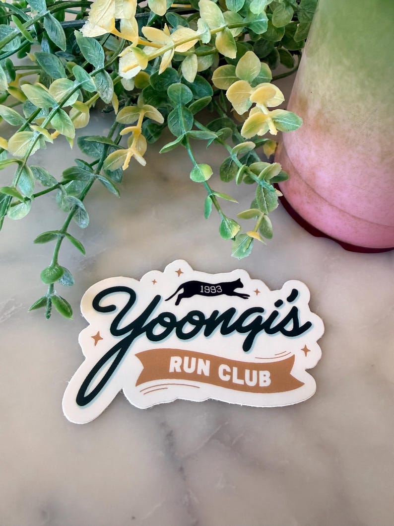 May include: A die-cut sticker featuring the black text Yoongi's in a cursive font, with the words RUN CLUB in white on a tan banner. Above the text is a black silhouette of a running cat with the number 1993, accented by small four-pointed stars.