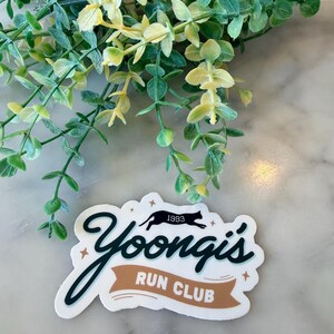 May include: A die-cut sticker featuring the text Yoongi's in dark green cursive script above a tan banner with the white text RUN CLUB. A black silhouette of a running cat with the year 1993 above it is centered between small gold stars.