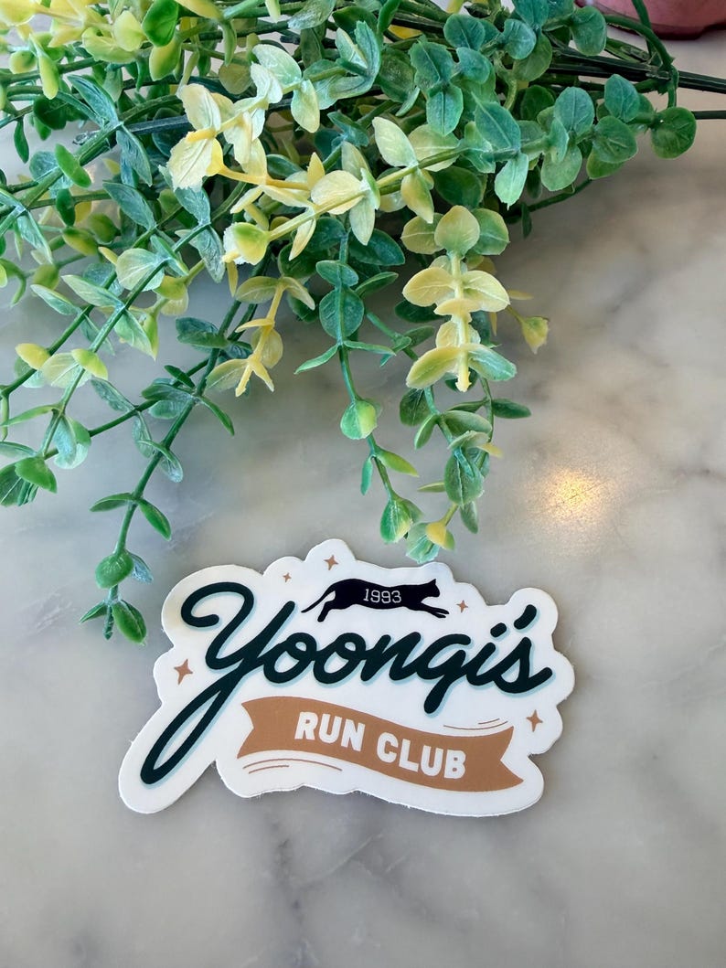 May include: A die-cut sticker featuring the text Yoongi's in dark green cursive script above a tan banner with the white text RUN CLUB. A black silhouette of a running cat with the year 1993 above it is centered between small gold stars.