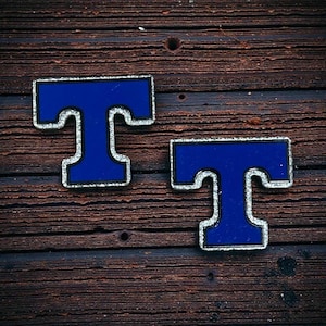 Puede incluir: Dos pines azules con la letra "T" y ribete plateado se muestran sobre una superficie de madera marrón oscura y envejecida. Los pines tienen un diseño de letra en bloque audaz, con un centro azul brillante y un contorno plateado.