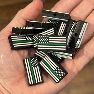May include: A pile of black, white and green silicone charms featuring the American flag with a thin green line. The charms have a hole for attaching to a keychain or other items.