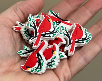 Può includere: Una collezione di ciondoli a forma di uccello cardinale rosso con rami neri e foglie verdi. I ciondoli sono bianchi ai bordi. Sono tenuti in una mano.