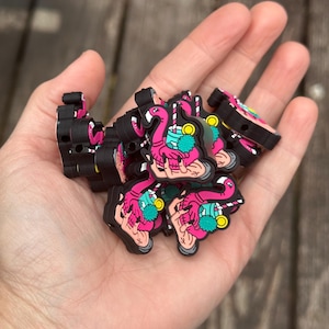 Op de afbeelding: Een handvol zwarte, roze en turquoise flamingo-bedels. Elke bedel toont een flamingo die een tropische drank met een roze rietje vasthoudt. De bedels zijn klein en hebben een 3D-ontwerp.