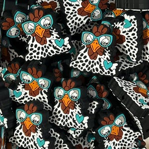 May include: A collection of silicone shoe charms, each shaped like a chicken head with a white and black spotted pattern. The chicken features a brown comb, an orange beak, teal-rimmed glasses, and a small teal heart accent on the side.