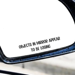 May include: A black car side mirror with the text "OBJECTS IN MIRROR APPEAR TO BE LOSING" in black, centered on the mirror's reflective surface. The mirror is set against a blurred outdoor background.