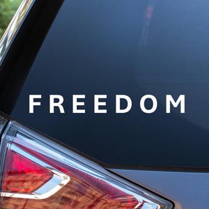 May include: A white vinyl decal displaying the word "FREEDOM" in a bold font is applied to the rear window of a car. The decal is centrally positioned and easily seen against the dark glass. The car's rear light is visible.