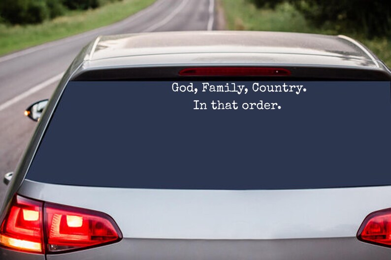 May include: Rear window of a silver car with a dark blue tint. White text reads "God, Family, Country. In that order." The car is on a road with a blurred background of trees and asphalt.