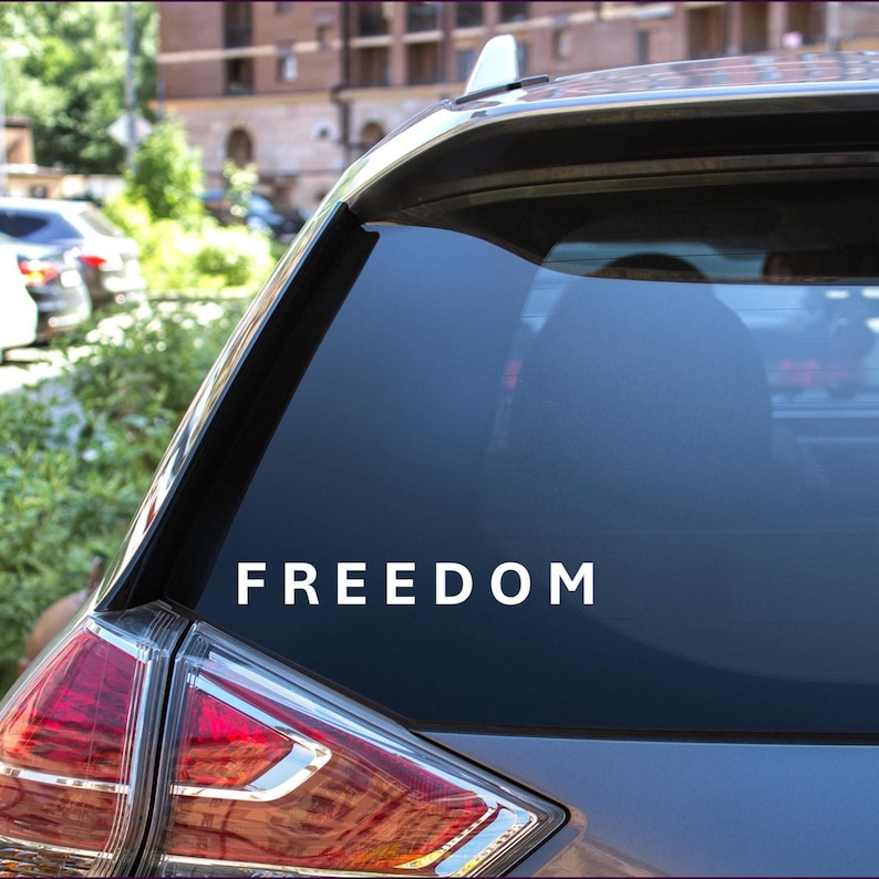 May include: A white vinyl decal displaying the word "FREEDOM" in a bold font is applied to the rear window of a car. The decal is centrally positioned and easily seen against the dark glass. The car's rear light is visible.