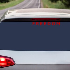 May include: Rear view of a silver car with a dark blue tinted rear window. The word "FREEDOM" is displayed in red lettering across the top of the window. The car is on a road with a green background.