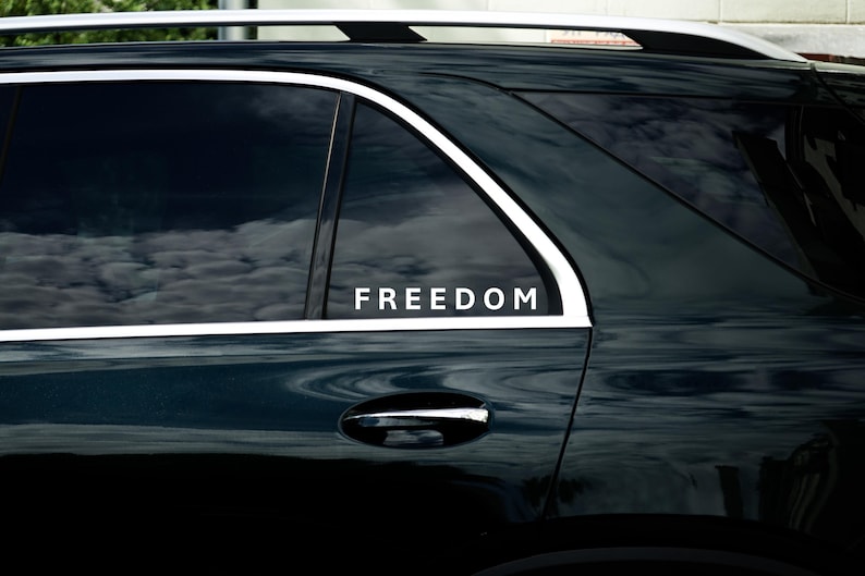May include: A dark green car window displaying the word "FREEDOM" in white. The window reflects a cloudy sky. The car features a silver roof rack and a chrome door handle.
