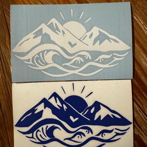 May include: Two vinyl stickers with a mountain range and ocean wave design. The top sticker is white on a light blue background. The bottom sticker is blue on a white background. Both stickers feature a sun and a heart.