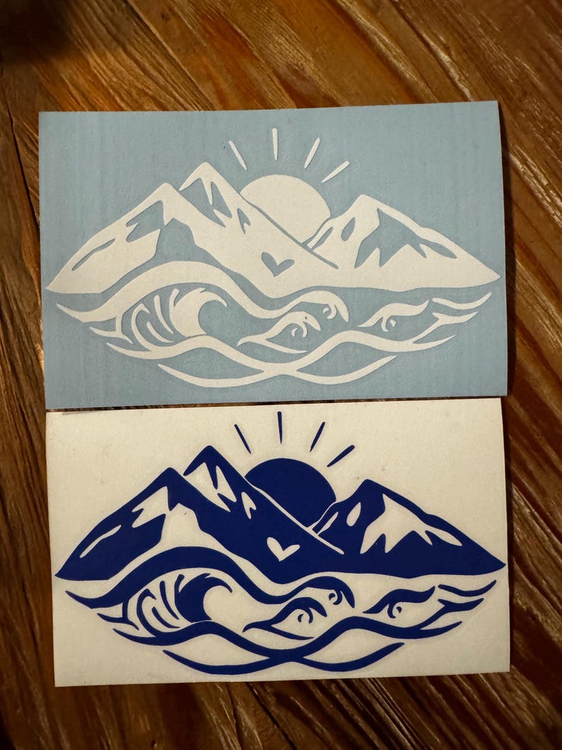May include: Two vinyl stickers with a mountain range and ocean wave design. The top sticker is white on a light blue background. The bottom sticker is blue on a white background. Both stickers feature a sun and a heart.