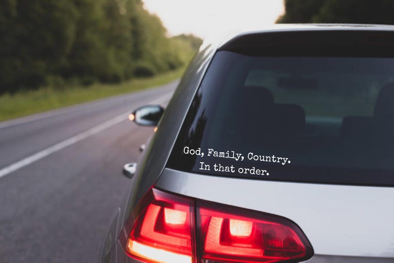 May include: A silver car's rear window features a white sticker with the words "God, Family, Country. In that order." The car is on a road with trees. The taillights are red.