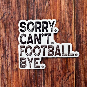 May include: A white sticker with black text that reads "SORRY. CAN'T. FOOTBALL. BYE." The sticker is on a wooden surface with vertical wood grain. The text has a distressed, vintage style.