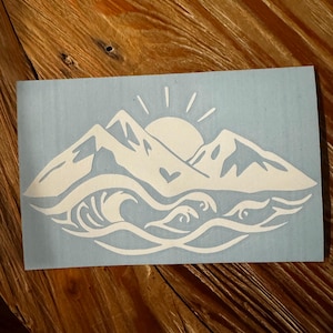 May include: White vinyl decal with a mountain range silhouette, a sun with rays, and a wave with a heart in the center. The design is on a light blue background.