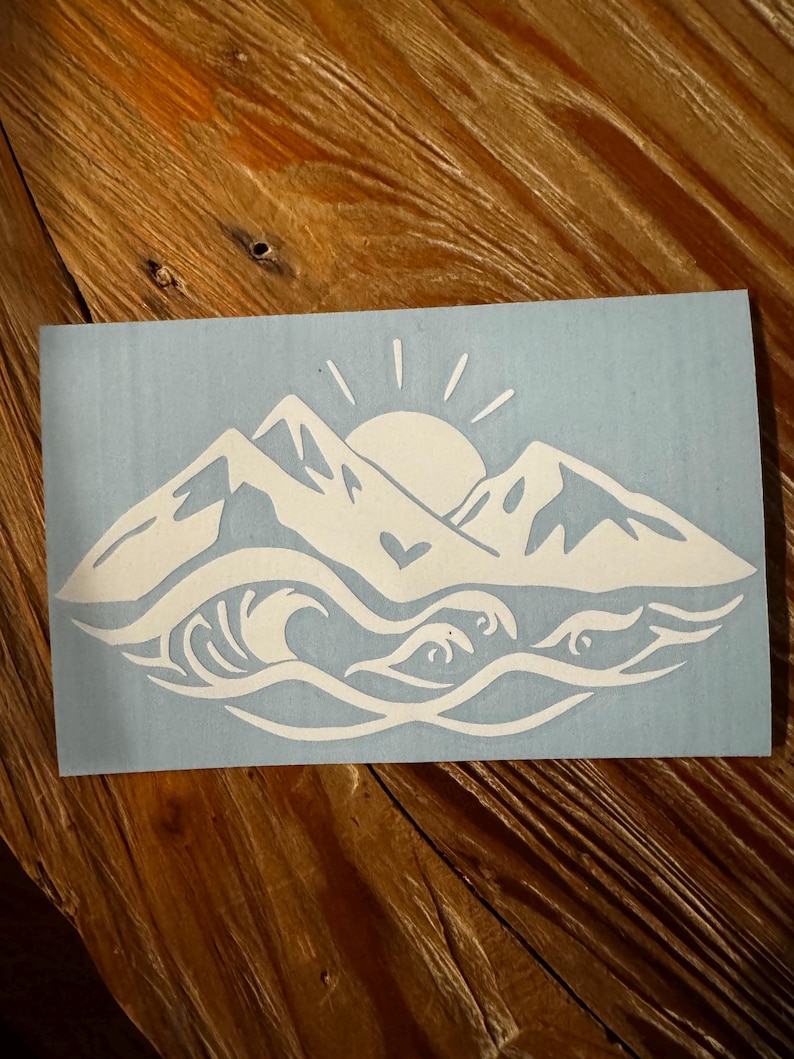May include: White vinyl decal with a mountain range silhouette, a sun with rays, and a wave with a heart in the center. The design is on a light blue background.