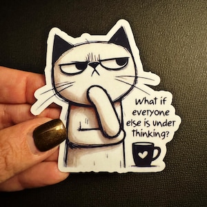 May include: A white sticker featuring a grumpy cat with black ears and eyebrows, pondering with its paw on its chin. The sticker includes the text "What if everyone else is under thinking?" and a small coffee cup.