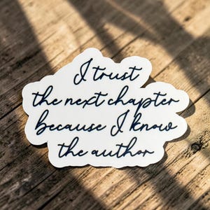 May include: A white die-cut sticker with a dark blue cursive script that reads, I trust the next chapter because I know the author. The sticker has an irregular, rounded edge shape and is displayed on a textured wooden surface with light shadows.