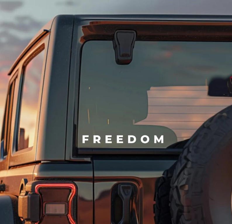 May include: The rear window of a black SUV features the white word "FREEDOM". The vehicle is parked outside, with a sunset visible through the window. A spare tyre is mounted on the back.