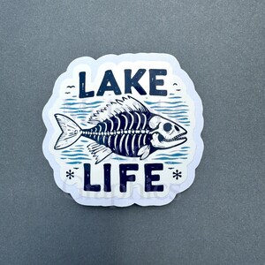 May include: A white sticker with a blue fish skeleton design. The fish is swimming through wavy lines. The text "LAKE LIFE" is written in blue letters above and below the fish.
