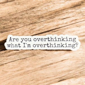 May include: A white sticker with black text that reads, "Are you overthinking what I'm overthinking?" The sticker is rectangular with rounded corners and is placed on a wooden surface.