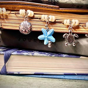 May include: A stack of journals with decorative charms hanging from the pages. Charms include a flip-flop, sand dollar, starfish, turtle, and dolphin. The charms are silver-toned with blue and white accents.